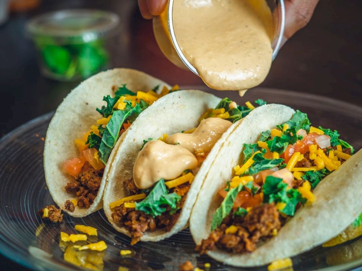 Three tacos filled with meat, cheese, lettuce, and tomato, topped with a creamy sauce being drizzled on.